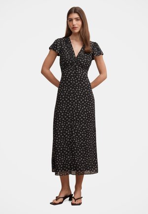 Frau trägt ein schwarzes Midi-Kleid mit kurzen Ärmeln und weißem Blumenmuster sowie schwarze Sandalen mit Absatz und steht vor einem schlichten Hintergrund.
