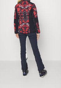 Skidjacka med ett rött och svart abstrakt mönster, svarta ärmar, kombinerad med marinblå byxor med vita sidostråk och dragkedjedetaljer.