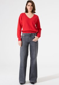 Pull à col en V texturé rouge associé à un jean large taille haute gris foncé. Le pantalon présente une fermeture à bouton à l'avant et des poches.