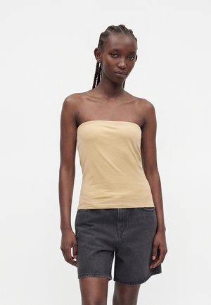 Donna con i capelli intrecciati che indossa un top beige senza spalline e pantaloncini in denim grigio scuro, in piedi su uno sfondo bianco uniforme.