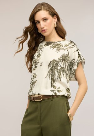 Frau mit langen, welligen Haaren, die eine weiße Bluse mit Blumenmuster, olivgrüne Hose, braunen Gürtel, goldene Ohrringe und ein goldenes Armband trägt, die Hände in den Hosentaschen.