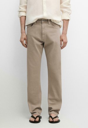 Mann trägt beige, gerade geschnittene Hose, cremefarbenes langärmeliges Hemd mit hochgekrempelten Ärmeln und schwarze Flip-Flops, steht vor einfachem Hintergrund.