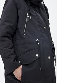 Czarna kurtka z kapturem, wyposażona w kieszeń na piersi na zamek, zatrzaski, ściągacz w pasie oraz kontrastujące srebrne elementy ozdobne o gładkiej fakturze.