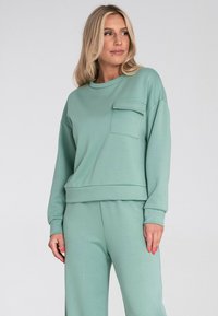 Jasnozielona bluza z długimi rękawami, okrągłym dekoltem i przednią kieszenią, w zestawie z dopasowanymi spodniami, wykonana z miękkiego materiału o gładkiej teksturze.