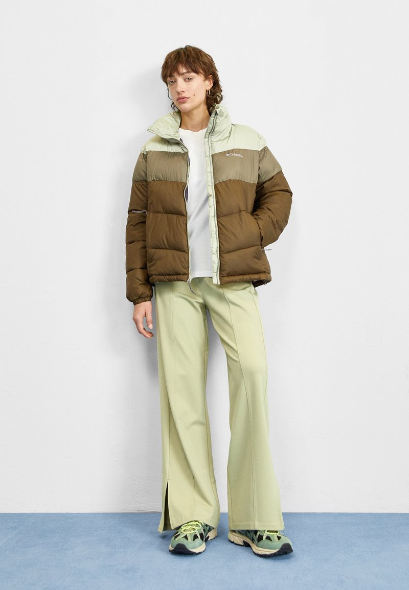 Chaqueta acolchada en verde oliva y menta clara, con cremallera frontal y cuello alto. Lleva unos pantalones verde claro y zapatillas con un diseño estampado.