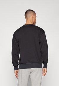 Homem com cabelo curto a usar um sweatshirt preto de mangas longas e calças de fato de treino cinza claro, de pé de costas contra um fundo simples.