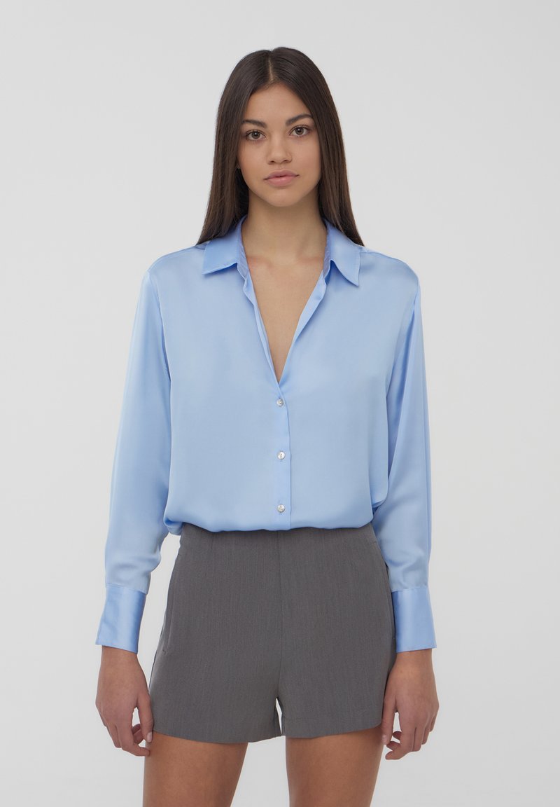 Camicia button-up in satin blu chiaro con scollatura a V e maniche lunghe, abbinata a pantaloni corti grigi a pieghe. Il tessuto appare liscio e lucido.
