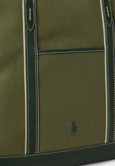 Πράσινη τσάντα καμβά με σκουρότερες πράσινες λεπτομέρειες, με μπεζ ραφές, μπροστινή τσέπη και διακριτικό λογότυπο ανάγλυφο στην πλευρά.