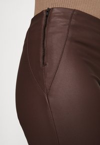 Gros plan sur un pantalon marron ajusté avec un zip à l'arrière, mettant en valeur des coutures texturées et un tissu au fini mat contre un haut beige.