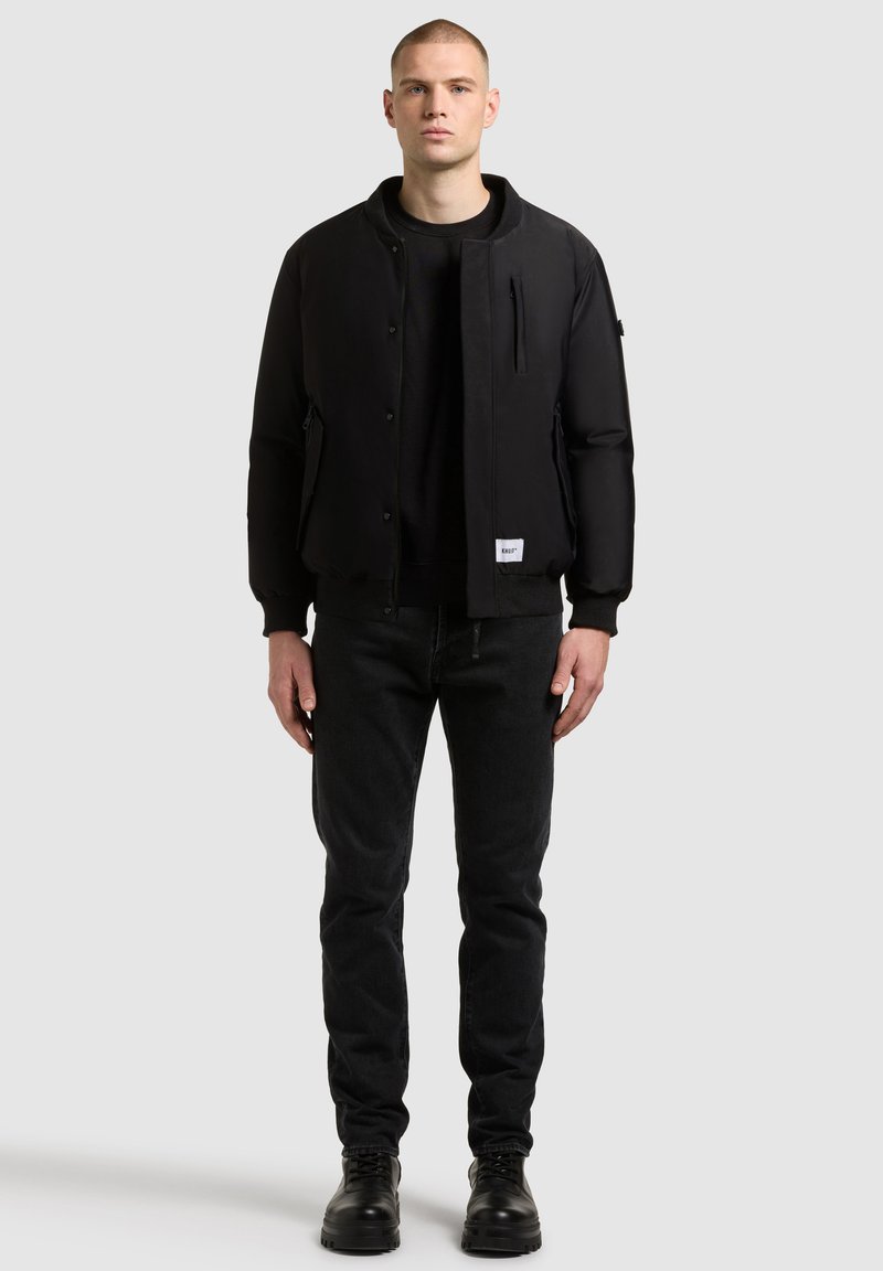 Czarna kurtka bombowiec z okrągłym dekoltem, przednią kieszenią na zamek, bocznymi kieszeniami i ściągaczami na mankietach, w zestawie z ciemnymi dżinsami i czarnymi butami.