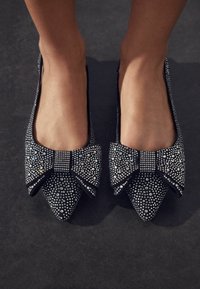 Juodos plokščios bateliai su smailiu nosiu, papuošti dideliu kaspinu ir metaliniais винтами tekstūrai. Turi lygią vidinę pamušalą.