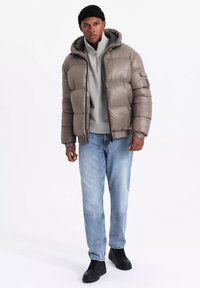 Giacca a vento color taupe, con cappuccio, chiusura con zip e polsini elasticizzati. Indossata sopra un pullover grigio e abbinata a jeans azzurri chiari e scarpe nere.