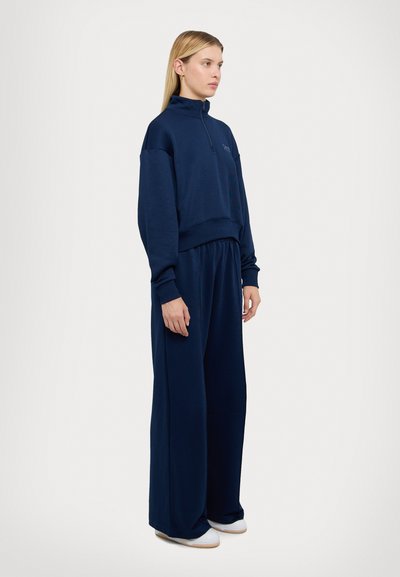 Pull à col zippé bleu marine avec une coupe décontractée, associé à un pantalon large de couleur et de texture assorties. Tissu doux, détails minimaux.