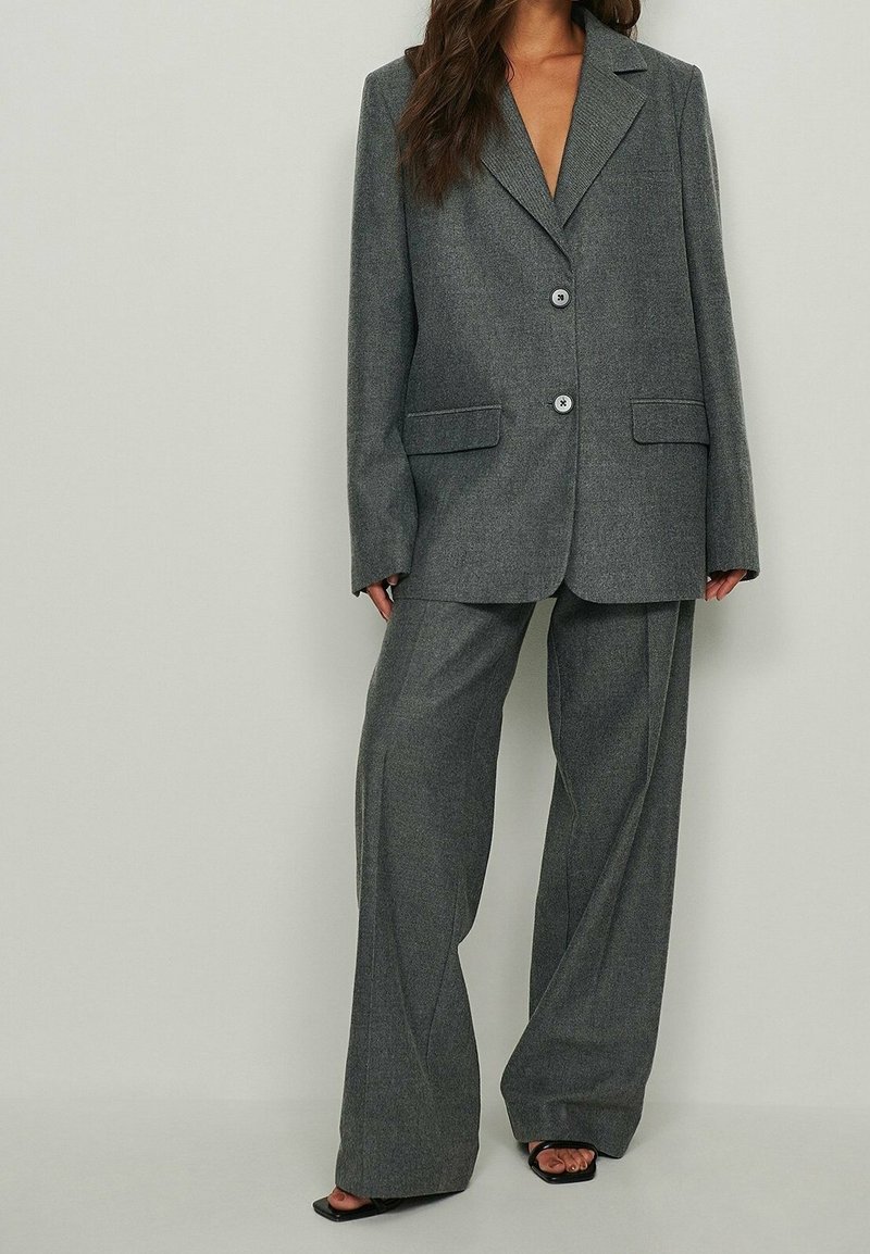 Femme portant un costume gris oversize avec un pantalon large et des talons ouverts noirs, debout devant un fond uni et clair.