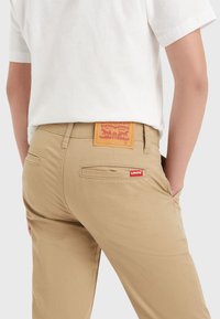 Pantaloni khaki dal tessuto liscio, con un'etichetta rossa Levi's sulla tasca posteriore e una patch in pelle beige con dettagli grafici rossi.