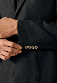 Dunkler, taillierter Blazer mit glatter Textur, vier runden Metallknöpfen am Ärmel und sichtbaren Nähten am Ärmel.