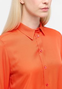 Orange, langærmet skjorte med klassisk krave og fem lyse knapper. Glat tekstur, tætsiddende design, tæt på billede af kraven og detaljerne.