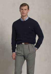 Marineblauwe trui met een ronde hals gedragen over een lichtblauw en wit gestreept shirt. Grijze geplooide broek en eenbruine riem. Soepele textuur.