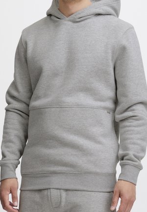 Homme portant un sweat à capuche gris clair uni à manches longues et un pantalon de survêtement gris assorti, debout devant un fond uni.