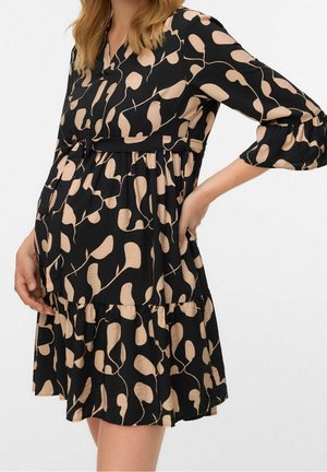 Kobieta ubrana w czarno-beżową sukienkę ciążową z wzorem, z rękawami do łokci, trzymająca brzuch i stojąca na białym tle.