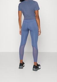 Blue athletic leggings featuring a mesh panel design and a stretch fabric. Paired with a grey fitted top. Black sports shoes visible.