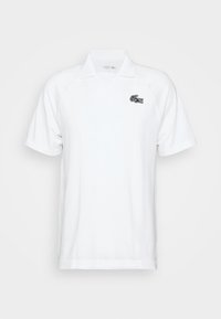 Bílá polo košile s krátkými raglánovými rukávy, měkkou texturou a černým logem Lacoste na levé straně hrudi. Klasický design límce.