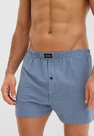 Torso masculin portant un boxer à rayures verticales bleues et blanches avec un bouton et une étiquette portant la mention "CASUAL." Mains détendues le long du corps.