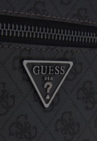Borsa nera texture con una placca logo triangolare argentata con "GUESS USA" e un punto interrogativo, sopra una chiusura a zip con denti metallici.