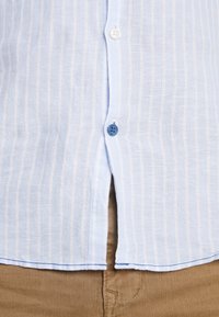 Chemise rayée bleu clair avec des lignes blanches ; présente un ourlet incurvé et des accents de boutons bleus ; fabriquée dans un tissu doux et texturé.