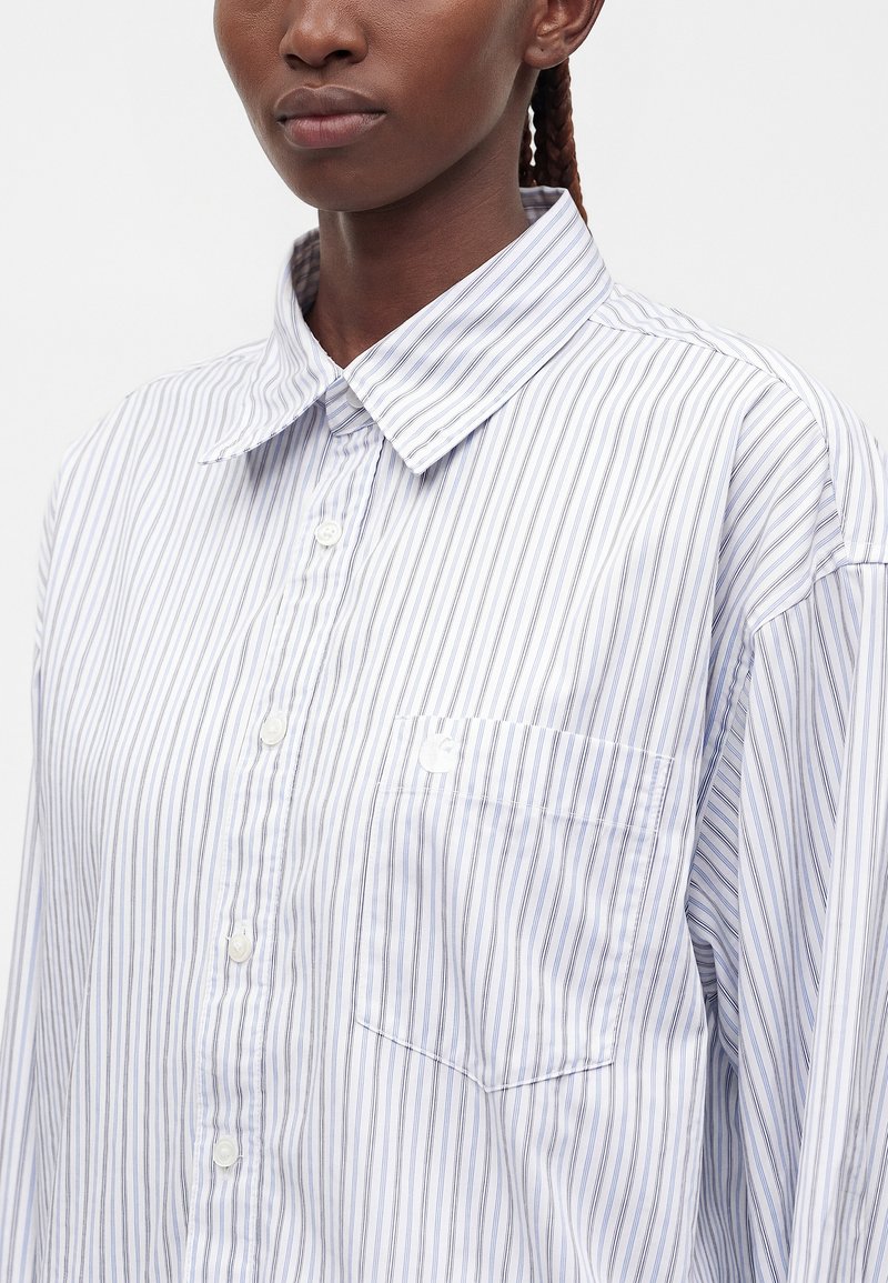 Donna che indossa una camicia bianca con sottili righe verticali blu e una tasca frontale, mostrata dal petto in su.