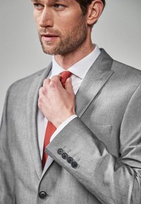 Grå jakkesætjakke med satinfinish, hvid skjorte og en orange slips. Funktioner inkluderer en notch-lapel og fem sorte knapper på ærmet.