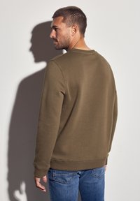 Mand i olivengrønt sweatshirt og blå jeans står med ryggen mod en hvid væg, mens han viser sin ryg og sideprofil med en skygge kastet bag sig.