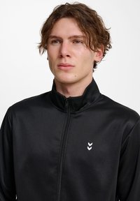 Sort zip-jakke med høj krave, der har en glat overflade og hvidt chevron-logo på venstre bryst.