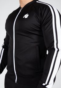 Veste de sport noire en matériau léger, avec une fermeture éclair blanche, deux poches latérales et des bandes blanches le long des manches. Logo gorille sur la poitrine.