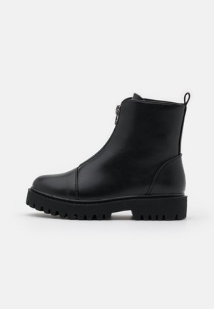 Bottine en cuir noir avec fermeture éclair à l'avant, semelle épaisse à crampons et languette à l'arrière, vue de profil sur fond blanc.