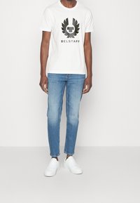 Camiseta blanca de algodón con un logo negro de Belstaff y diseño de alas, combinada con jeans de mezclilla azul claro y zapatillas blancas.
