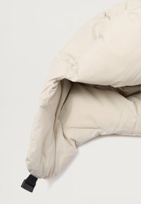 Piumone imbottito di crema con una texture liscia, caratterizzato da un design strutturato e una tracolla nera regolabile per la sicura.