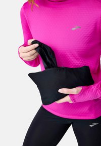 Haut à manches longues rose vif en tissu texturé, main tenant un pouch noir contenant un objet noir, sur fond blanc.