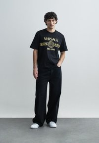 Black cotton T-shirt with gold "VERSACE MILANO" print and Greek key pattern. Paired with loose-fitting black pants and white sneakers.
