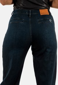 Pantalones de denim azul oscuro con corte recto, que presentan una parche de cuero marrón etiquetado "PLEASE" y dos bolsillos traseros con un detalle de corazón.