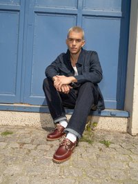 Chaussures en cuir marron avec semelles en caoutchouc, lacées de blanc, portées avec un jean bleu et une veste en denim foncé, assises contre une porte bleue.