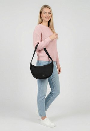 Sieviete rozā džemperī, gaiši zilās džinsās un baltos sporta apavos nes melnu pusmēness formas plecu somu, smaidot un stāvot pret vienmērīgu fona virsmu.