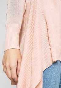 Ljust rosa ribbat stickat tröja med genomskinliga ärmar, har en svävande, asymmetrisk nederkant och texturerad tyg. Handen placerad nära nederkanten.