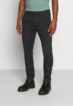 Pantalons fuselés noirs en coton avec une texture lisse, dotés d'une fermeture à bouton à l'avant et de poches latérales. Porté avec des bottes noires.