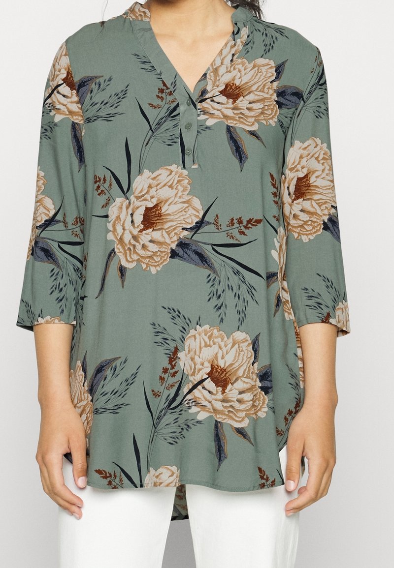 Femme portant une tunique verte avec un grand imprimé floral beige, manches trois-quarts, col en V boutonné, associée à un pantalon blanc.