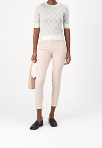 Helle rosa Skinny Jeans, kombiniert mit einem strukturierten weißen Kurzarm-Top mit einem Diamantmuster. Schwarze Slipper und eine kleine beigefarbene Handtasche runden den Look ab.