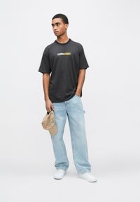 Μαύρο κοντομάνικο μπλουζάκι με κίτρινο και λευκό λογότυπο, συνδυασμένο με ανοιχτό μπλε φαρδιά τζιν και λευκά αθλητικά παπούτσια. Κρατάει μπεζ καπέλο.