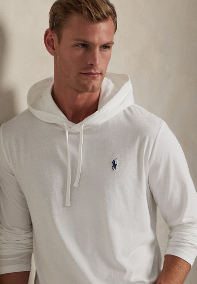 Mladić svijetlosmeđe kose koji nosi bijelu hoodicu s malim tamnoplavošim logom igrača poloa na lijevoj strani prsa.