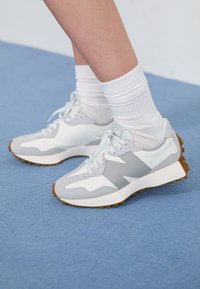 Chaussures de sport gris clair avec des accents blancs et une semelle en caoutchouc, présentant un mélange de matériaux, dont du mesh et du daim. Portées avec des chaussettes blanches.