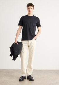 Black cotton T-shirt paired with light beige trousers. Model holds a black sweatshirt. Black loafers complete the outfit. Smooth textures.